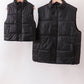 Two black puffer vests hanging on a rack against a white background